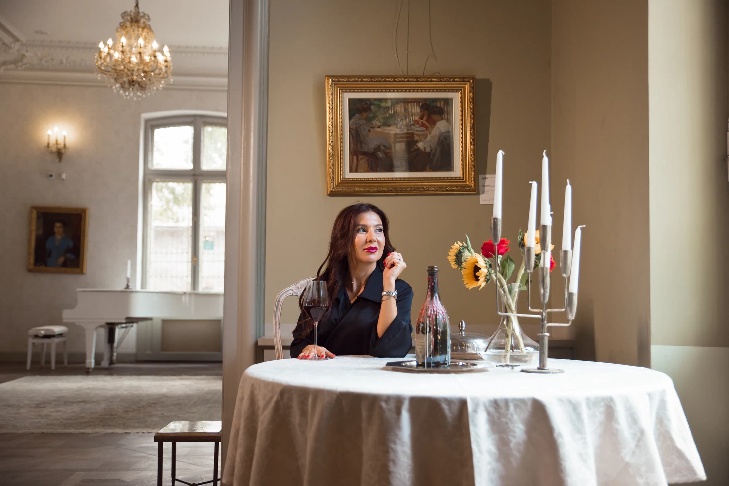 Roxana așezată la o masă rotundă cu lumânări și flori, sub un candelabru