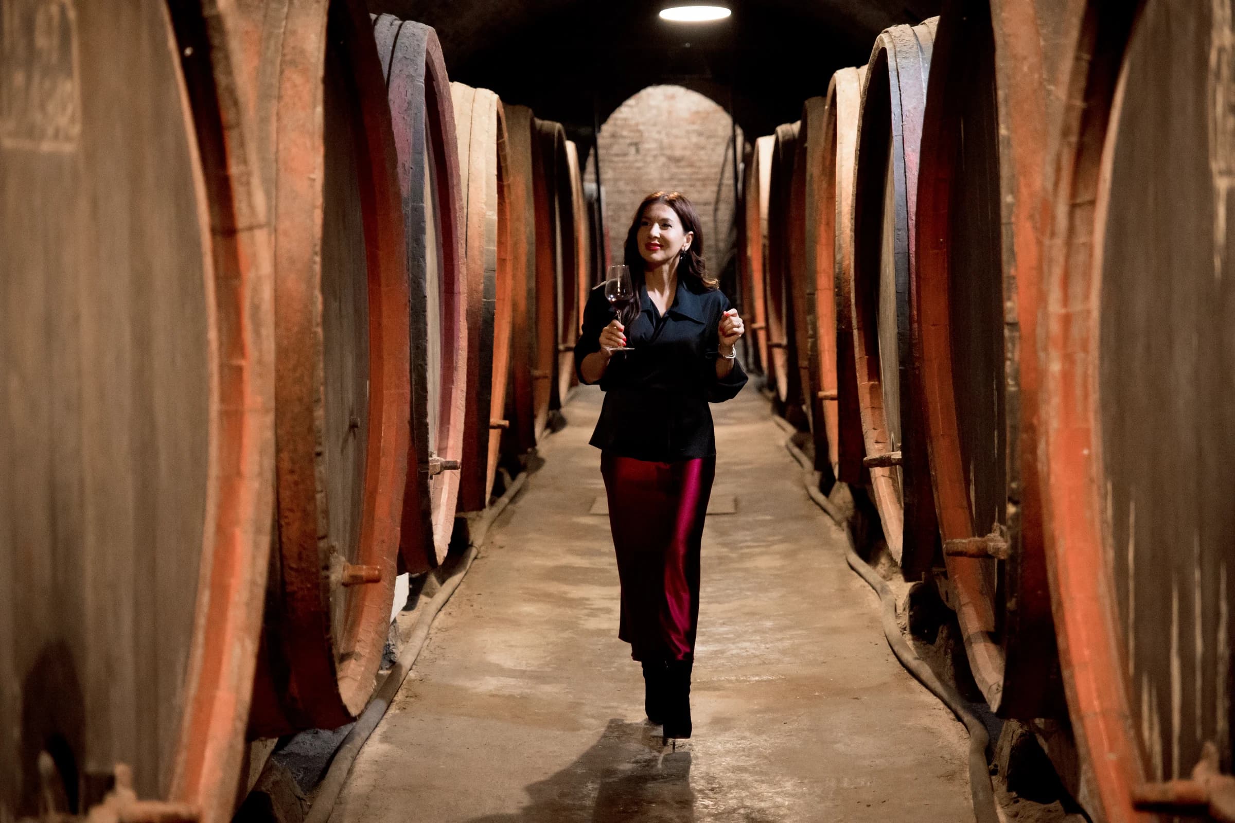 Roxana merge cu un pahar de vin printr-un coridor de butoaie mari de lemn